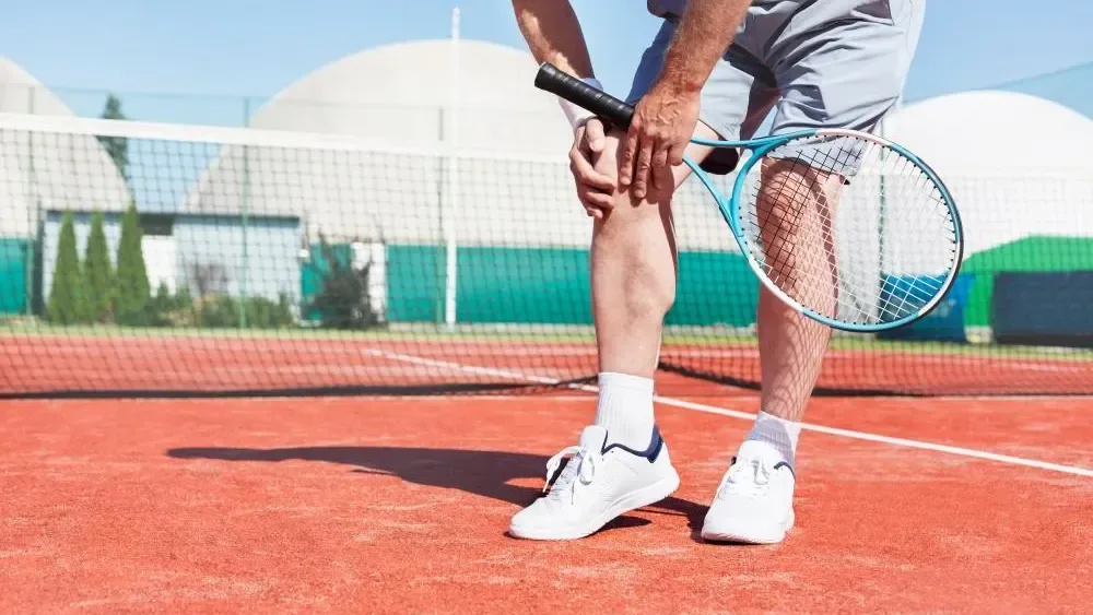 Tennis player on a red clay court holding his knee in pain while gripping his racket