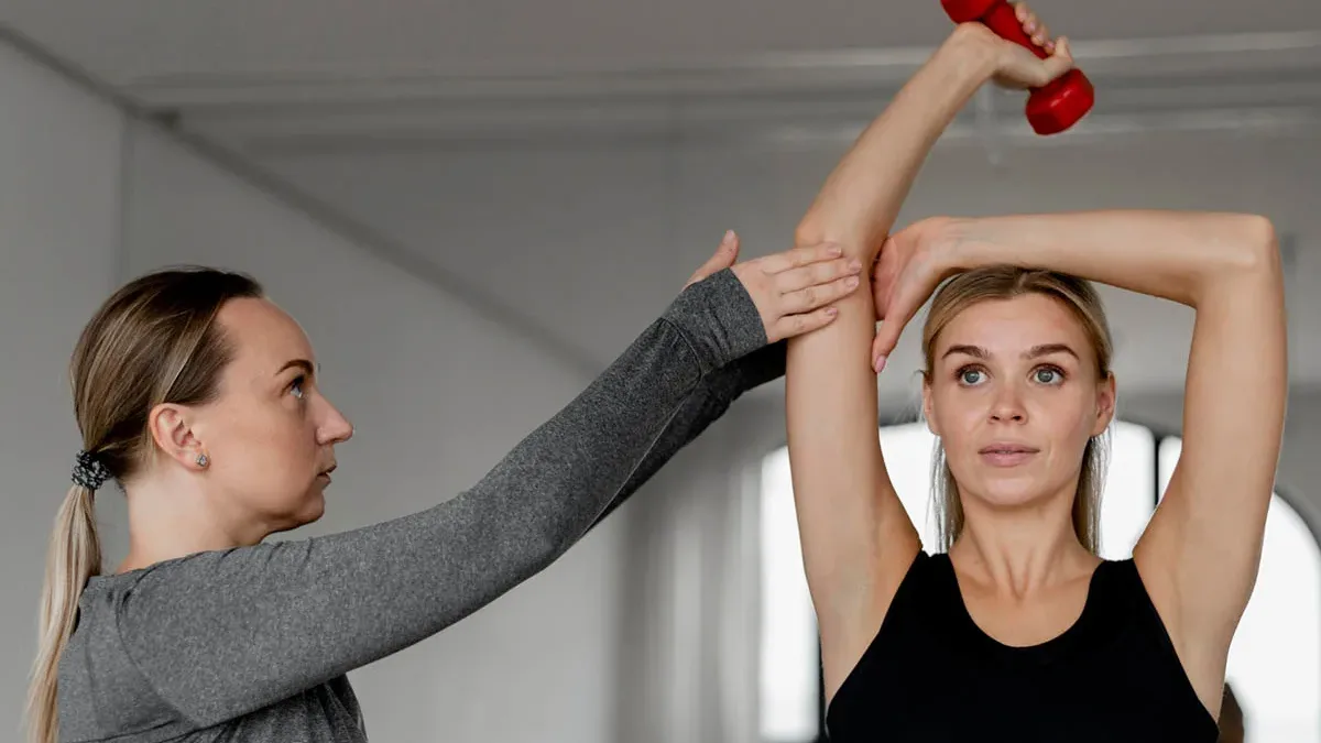 Physical therapist assisting a woman with an overhead shoulder stretch while she holds a dumbbell