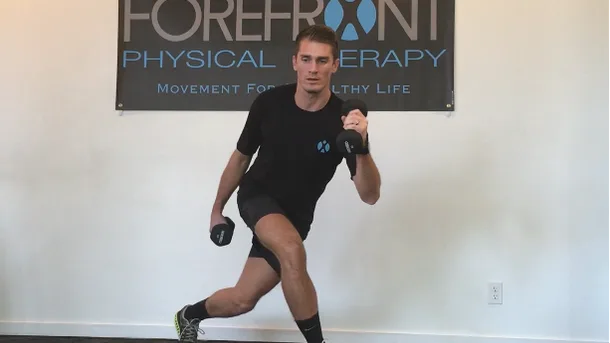 A man performing a walking lunge with dumbbells in front of a Forefront Physical Therapy banner