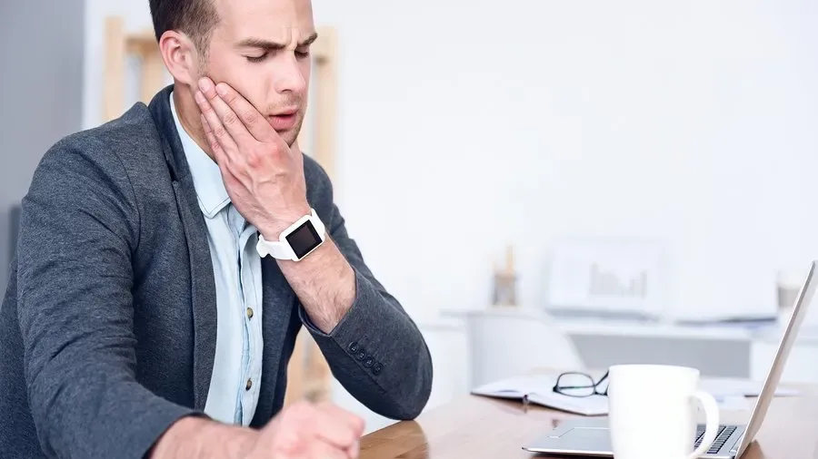 Man sitting at a desk holding his jaw in discomfort while working on a laptop