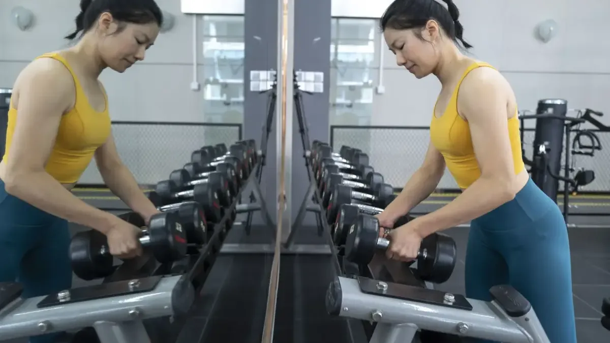 Woman lifting a dumbbell at the gym, reflected in a mirror alongside a rack of weights